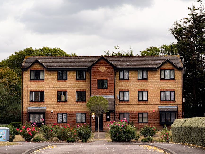 A three-story brick apartment building with a dark tiled roof, surrounded by neatly maintained garden beds filled with pink roses and green shrubs. The front garden features a small, well-trimmed tree